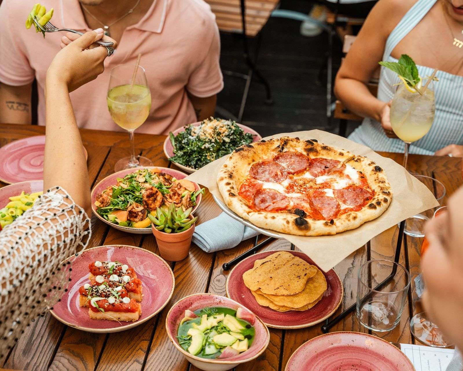 An overhead brunch spread of pizza, salads, and cocktails on pink stoneware at Odd Sister